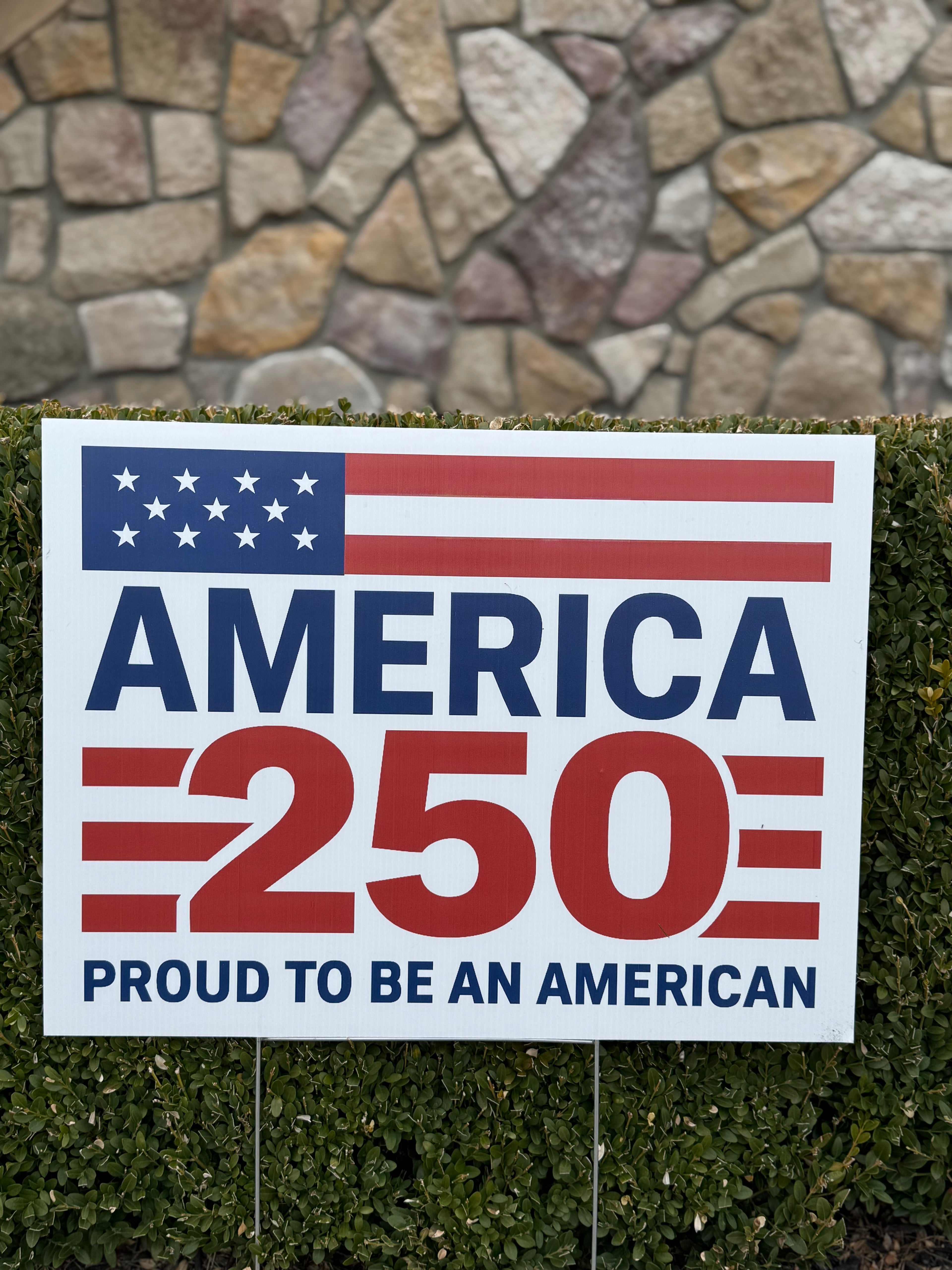 Bulk America 250 sign with American flag design and text 'America 250 Proud to be an American' against a stone wall background.