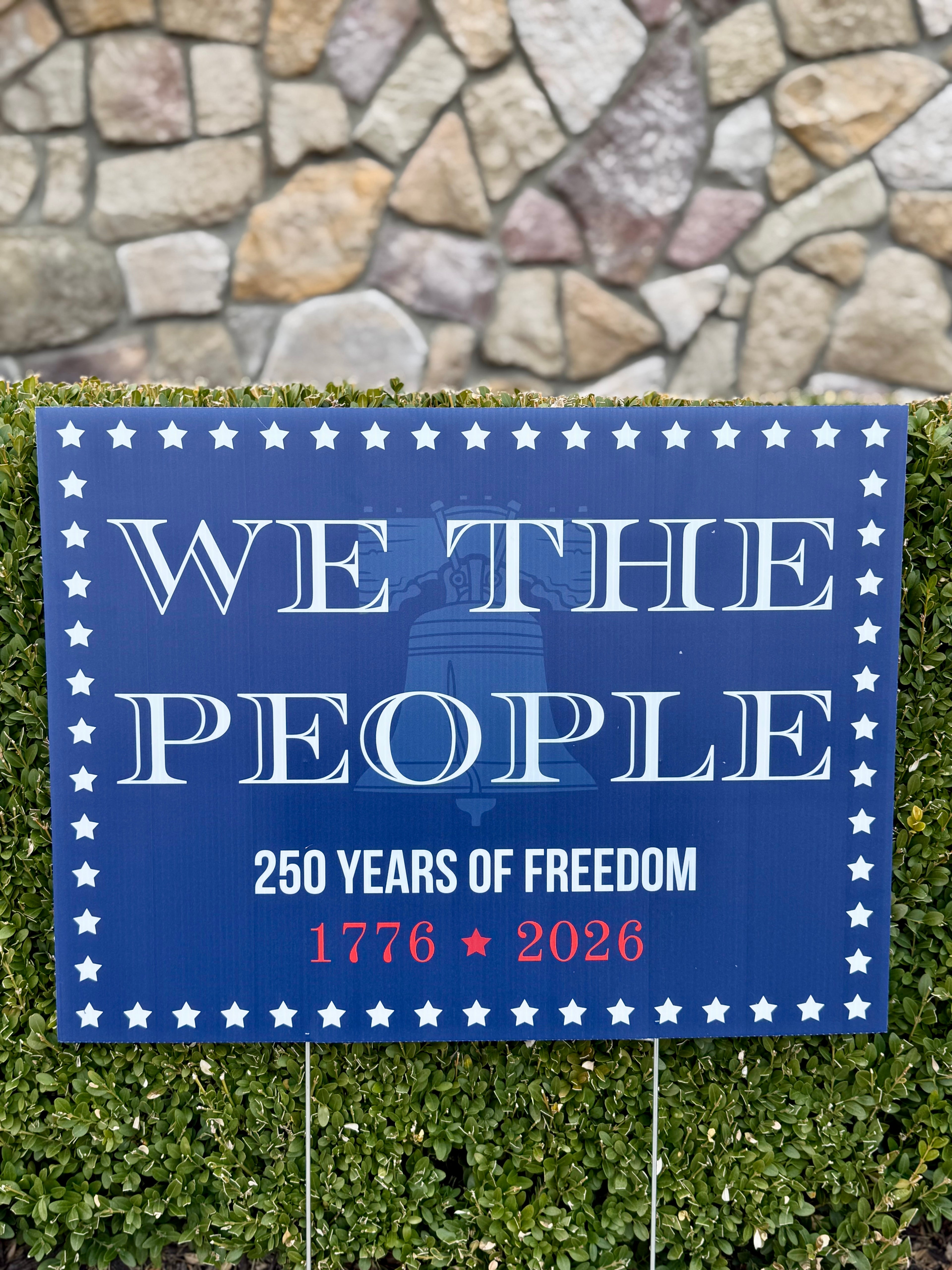 Bulk. A blue commemorative yard sign featuring the text "WE THE PEOPLE" in large white serif font above a faint outline of the Liberty Bell. Below it reads "250 YEARS OF FREEDOM" and "1776 ★ 2026" in red and white. The sign is bordered by small white stars and is displayed in front of a green hedge and a stone wall.
