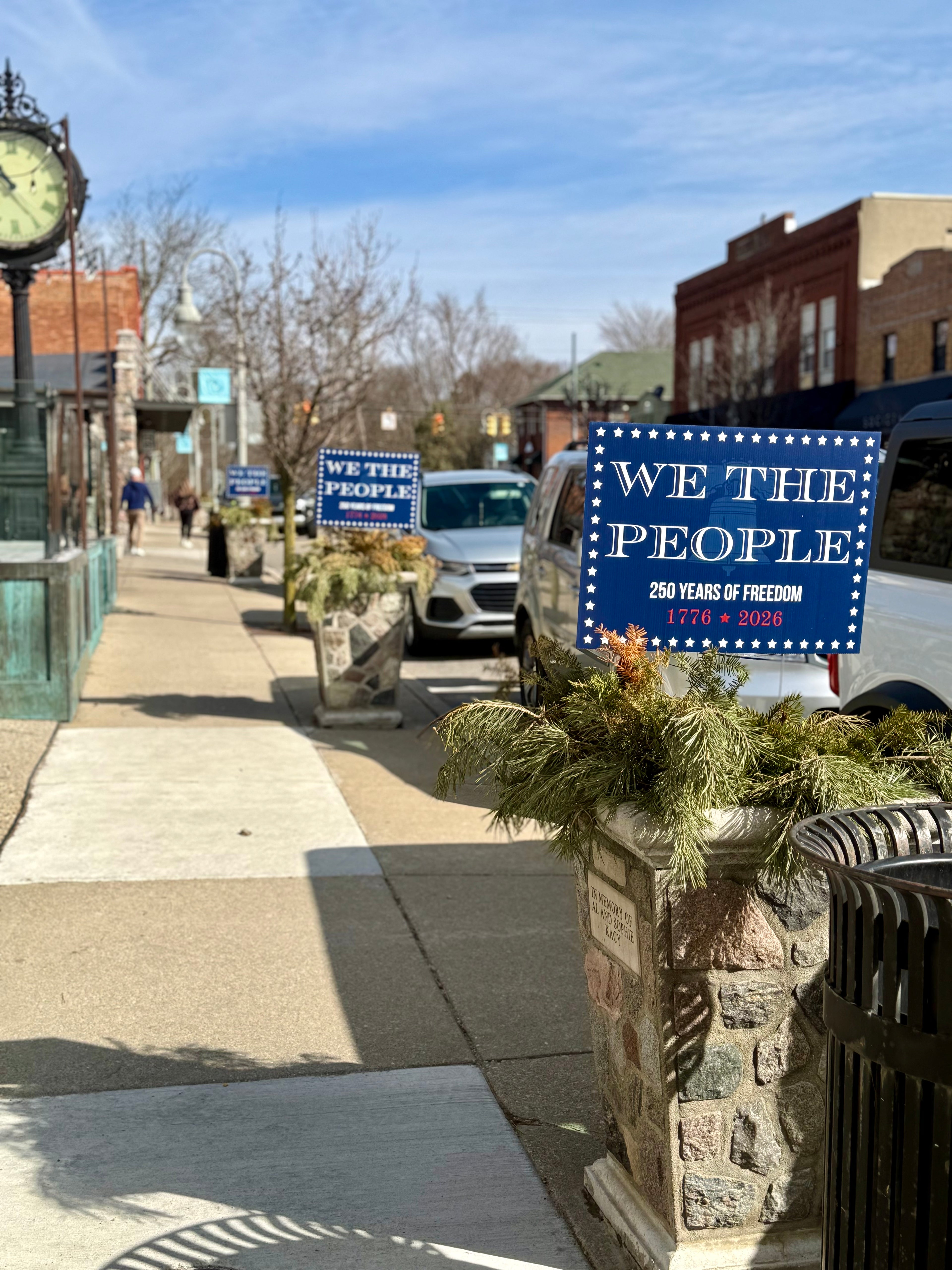 Decorative signs on a street corner with 'We the People' theme, featuring a clock and buildings in the background.
