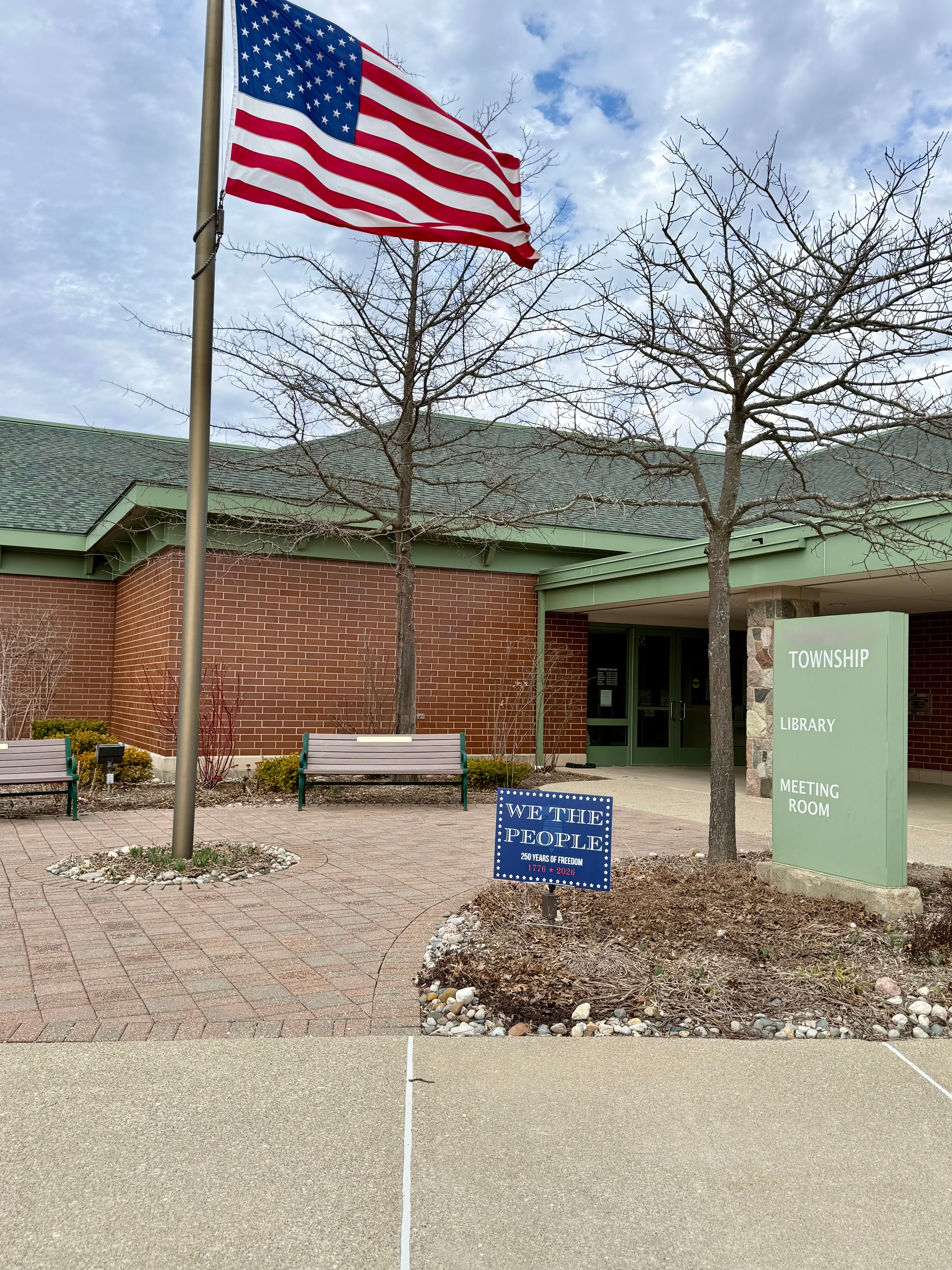 We The People Yard Sign at Township library with American flag, bench, and signs in front and bulk